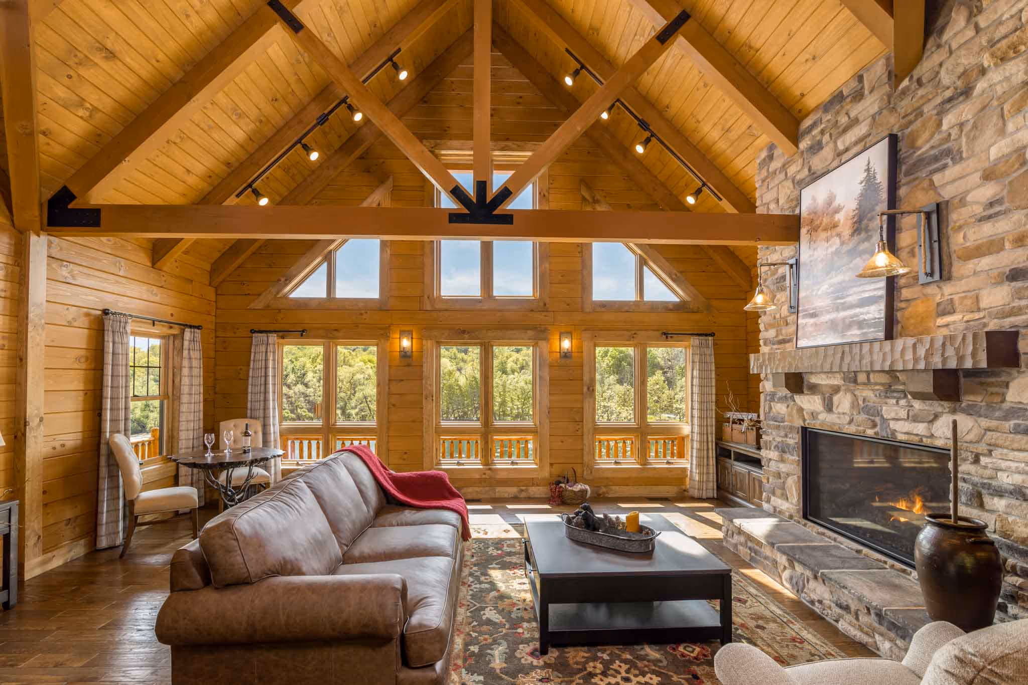 Interior of a log cabin living room featuring wooden beams, a striped couch, a fireplace, and large windows. A rocking chair and a round rug add warmth to the space. The room is well-lit with natural light.