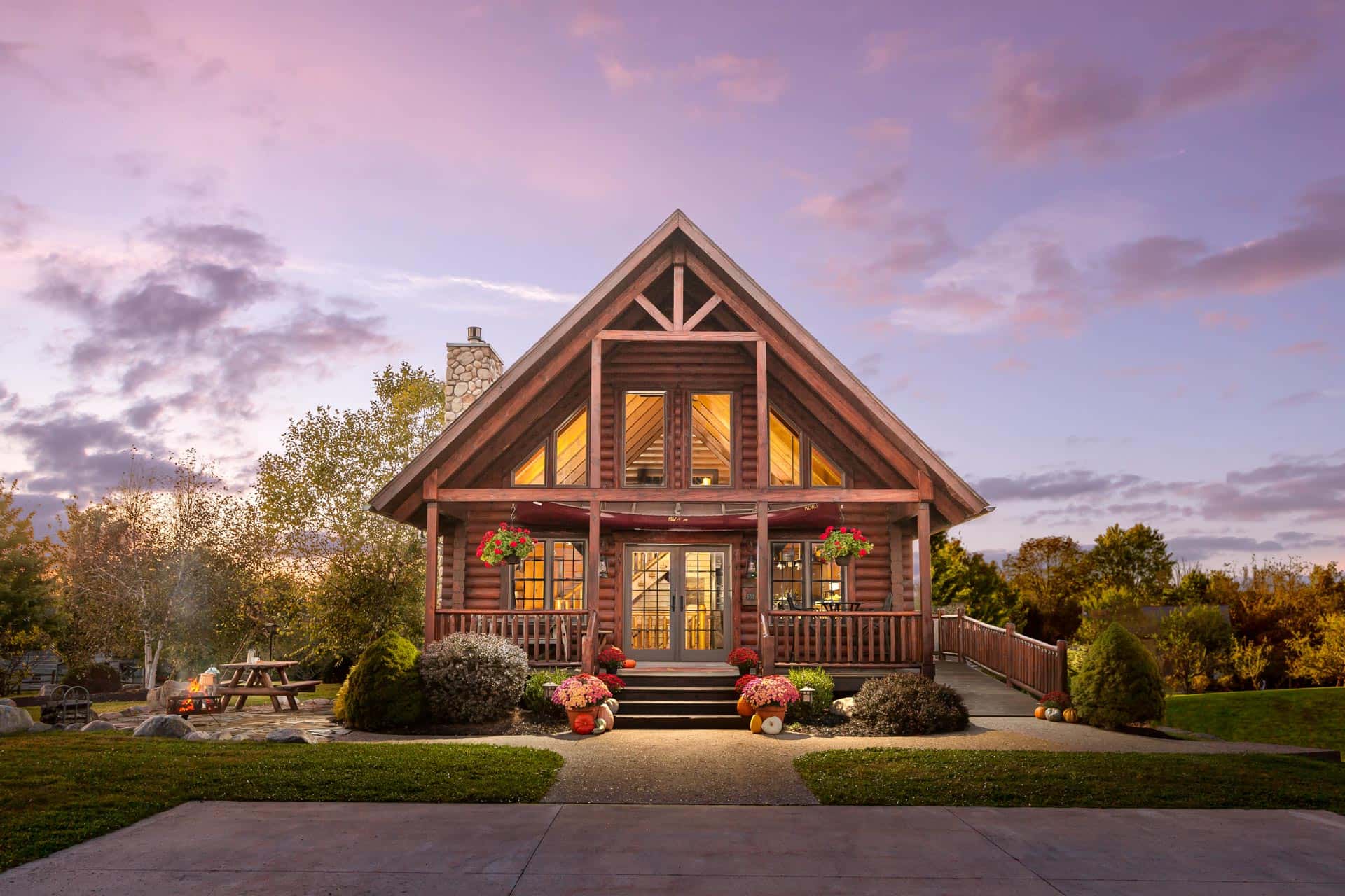 A modern log cabin surrounded by trees, featuring a large deck and stone accents. The structure has large windows, a peaked roof, and a stone chimney, set on a grassy area.
