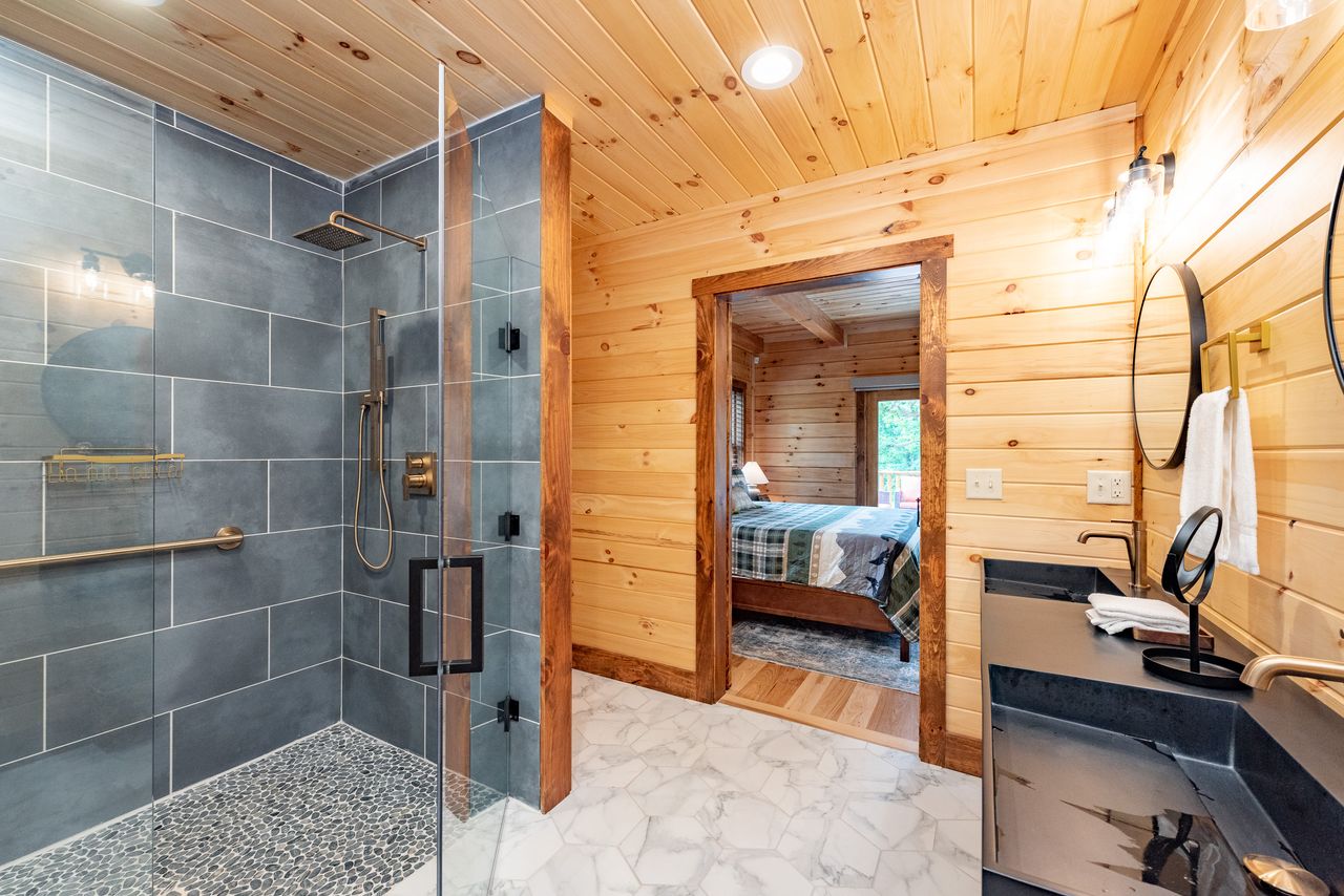 A modern bathroom featuring a walk-in shower with gray tiles, a glass enclosure, and a pebble floor. A black vanity with a round mirror is on the right, and a wooden door leads to a bedroom in the background.
