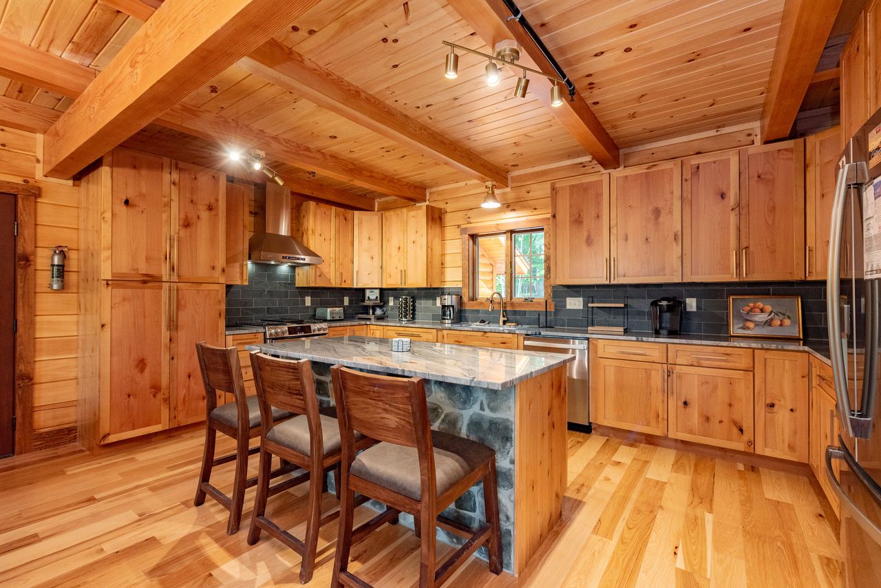 A spacious kitchen with wooden cabinetry, a large stone countertop island, and bar stools. The ceiling features exposed wooden beams, and large windows provide natural light. Appliances are in stainless steel, enhancing the modern look.