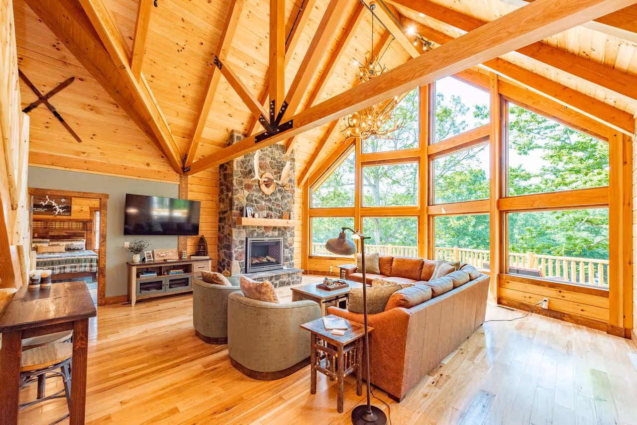 A cozy living room with a high wooden ceiling and large windows allowing natural light. The space features a stone fireplace, a comfortable sofa, and a side table with a lamp. Wooden flooring complements the rustic decor.