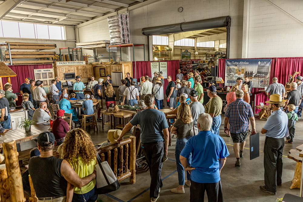 A busy indoor event showcasing log homes and custom homes, with many attendees viewing displays of log cabin kits and Amish homes. People are gathered around tables featuring various wooden products.