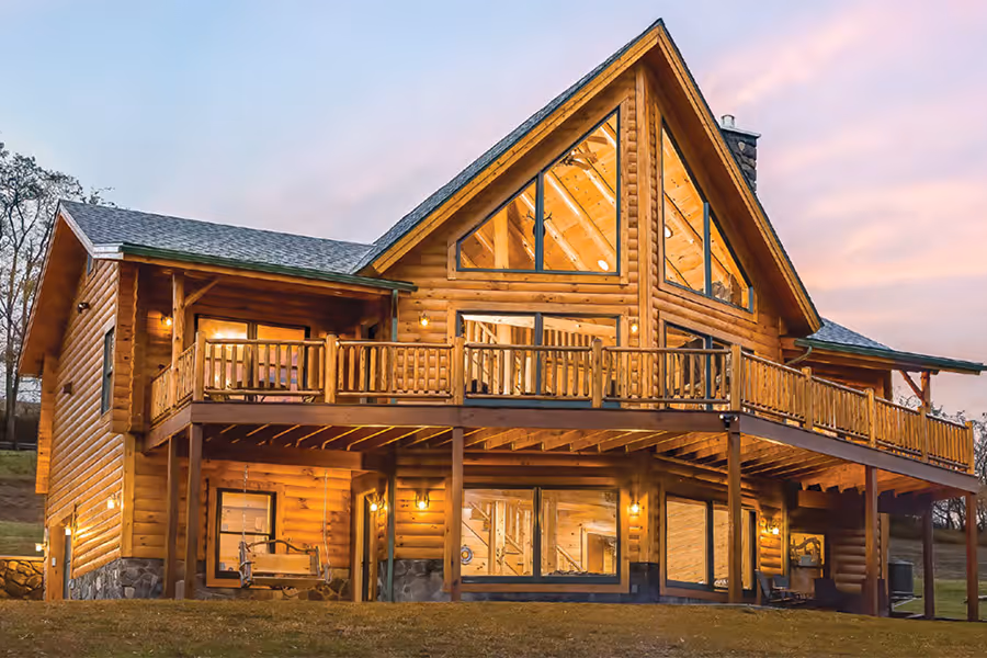 A large wooden log cabin with multiple balconies and large windows, set against a sunset sky. The structure features a steeply pitched roof and a warm, welcoming exterior. Surrounding greenery enhances the rustic charm of the home.