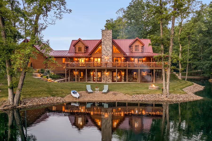 A beautiful wooden cabin with a red metal roof, situated by a serene lake. The two-story structure has large decks and windows reflecting in the calm water. Two chairs and a small boat are visible on the grassy bank, surrounded by lush trees.