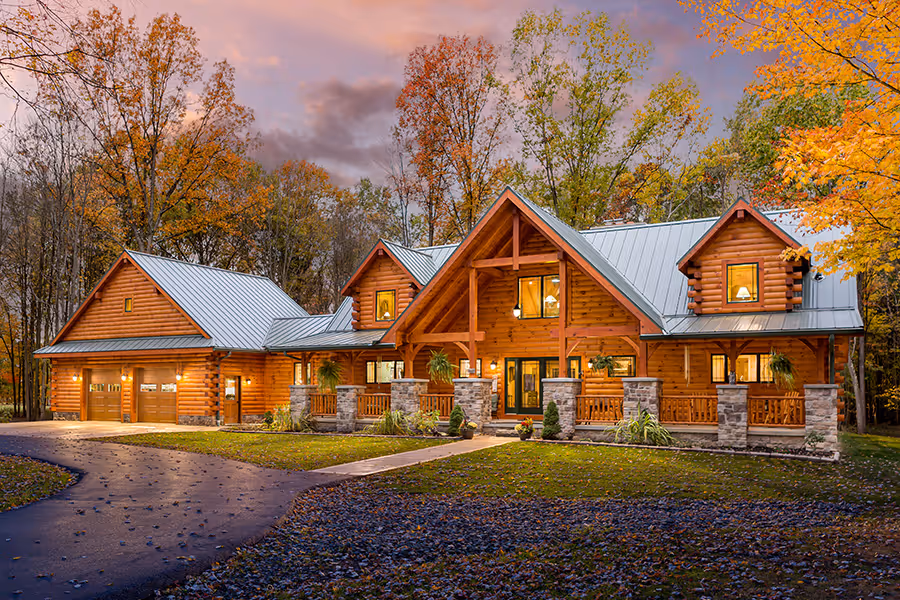 A beautiful log cabin surrounded by vibrant autumn foliage, featuring a stone and wood exterior, large windows, and a sloped metal roof. A winding driveway leads to the charming entrance, creating a picturesque setting.