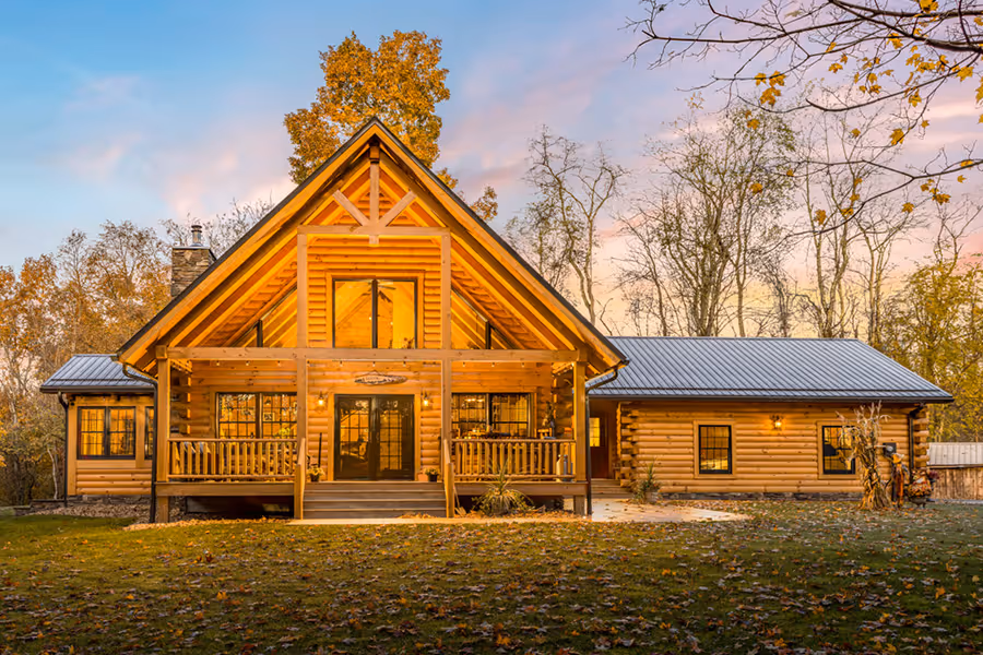 A charming log cabin with a peaked roof and large front porch, surrounded by trees and grass. Soft evening light illuminates the wooden structure, creating a cozy atmosphere.