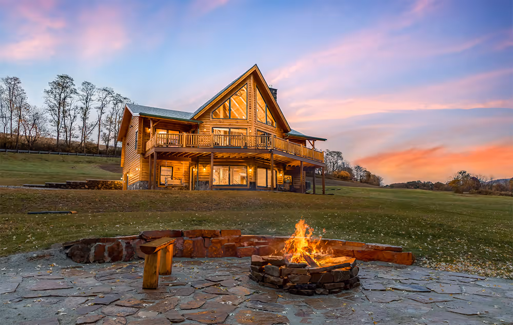 A cozy wooden cabin with large balconies, situated in a lush green landscape at sunset. A stone fire pit with a crackling fire is in the foreground, surrounded by a stone circle. The sky is painted in warm sunset hues.