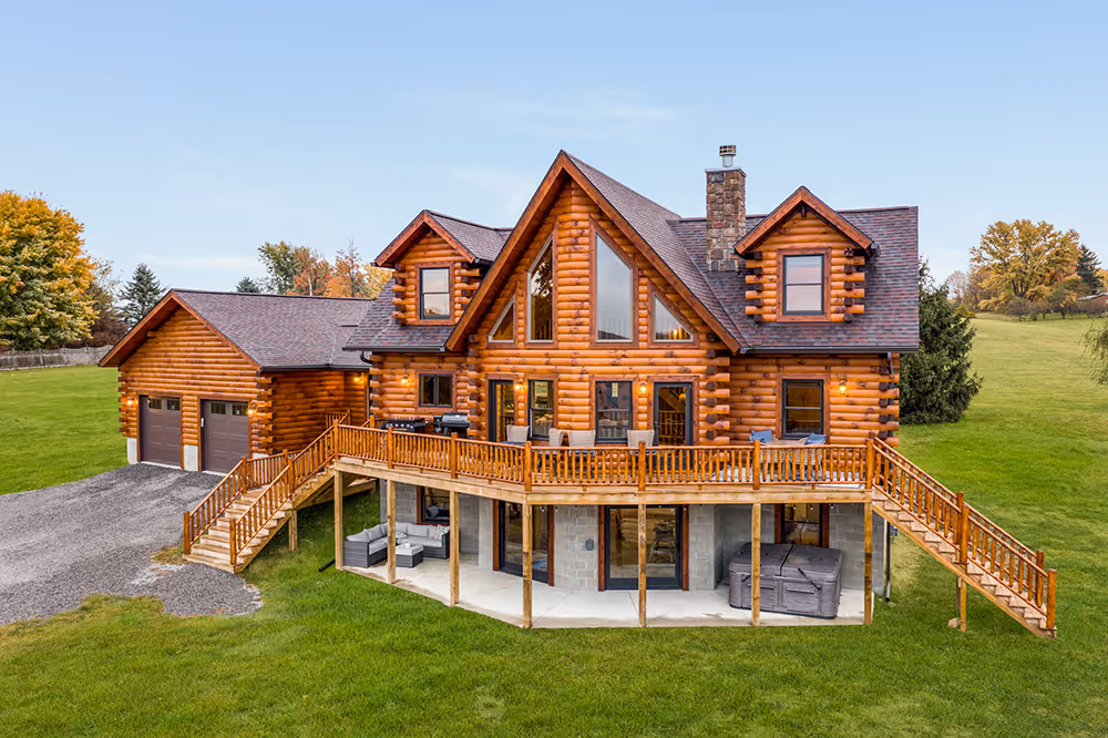 A spacious log cabin with a wooden deck, large windows, and a welcoming entrance surrounded by a green lawn and trees. The cabin features a garage and has a rustic charm, ideal for a cozy retreat.