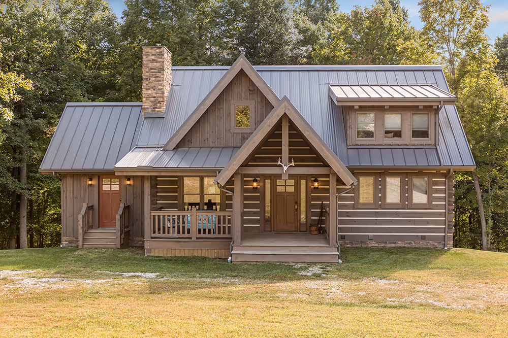 A rustic wooden cabin featuring a triangular roof, stone chimney, and front porch surrounded by trees. The cabin's exterior showcases a blend of logs and modern materials, set in a natural landscape with a green lawn.