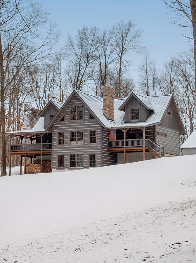 A large log cabin home surrounded by snow-covered trees, featuring multiple gable roofs, wide windows, and a front porch. Snow blankets the ground in front of the house, creating a serene winter landscape.