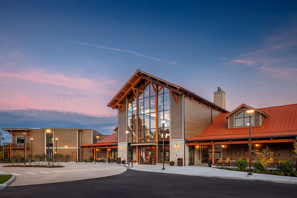 A modern lodge-style building with large glass windows and a red sloped roof, surrounded by landscaped grounds. The sky showcases a gradient of orange and purple hues during sunset, hinting at a welcoming atmosphere.