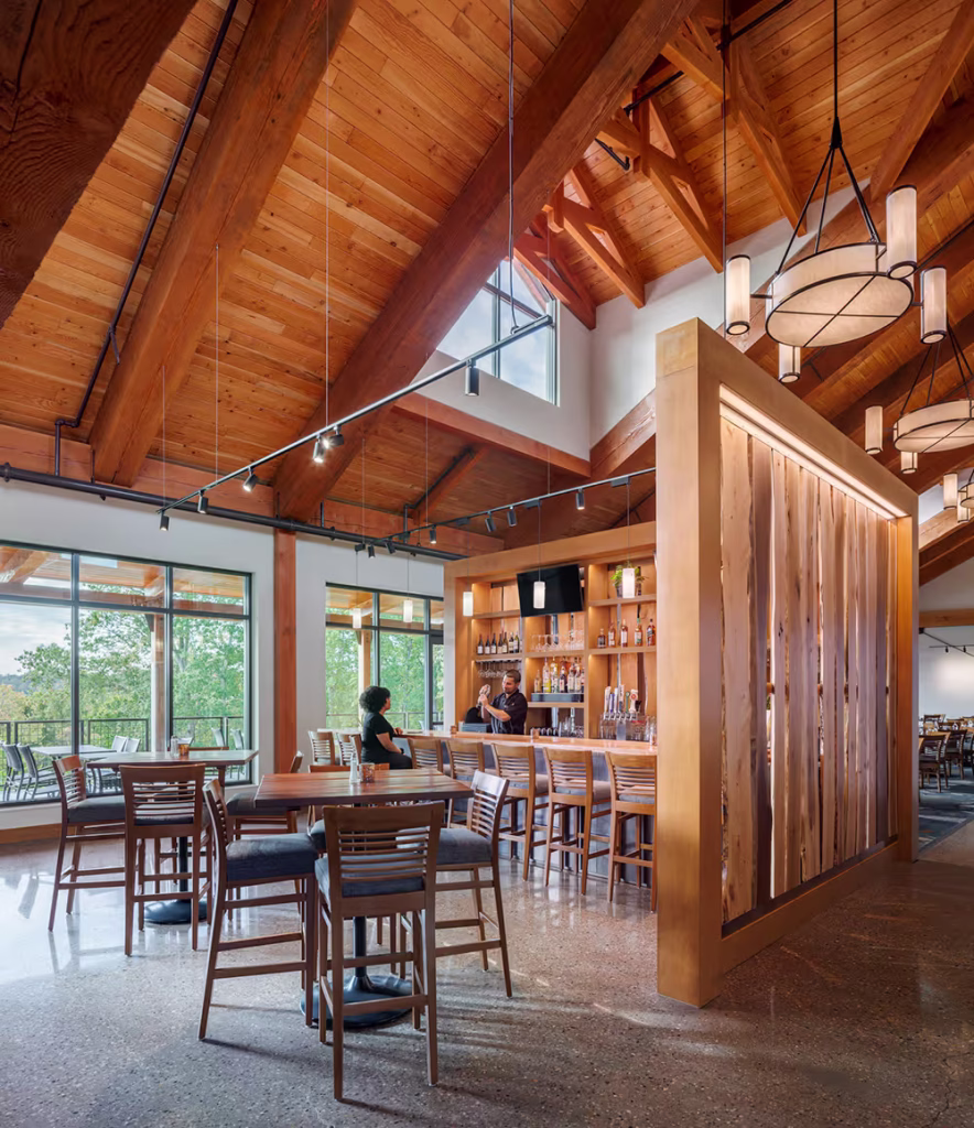 A modern restaurant interior featuring wooden beams and large windows. Tables with chairs are arranged inside, while two staff members converse near a wooden bar area displaying liquor bottles.
