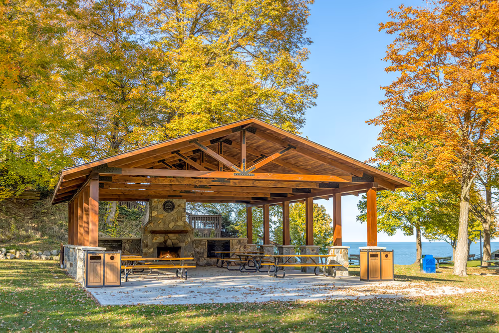 A rustic pavilion with a wooden roof and stone fireplace surrounded by green grass and colorful autumn trees, overlooking a serene lake with blue skies in the background.