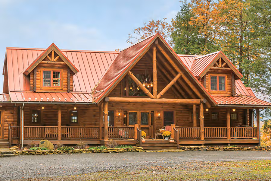 A charming log cabin with a red metal roof and wooden porch, surrounded by trees and fall foliage. The two-story structure features intricate woodwork and large windows, creating a warm and inviting atmosphere.