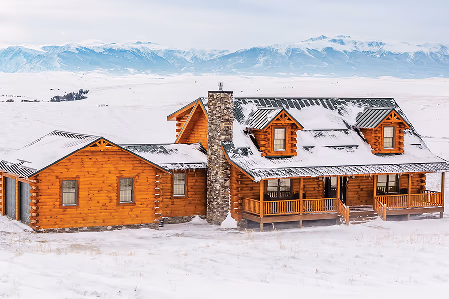 A charming wooden cabin with a stone chimney, surrounded by pristine snow and mountains in the background. The cabin features a sloped roof and a warm, inviting porch. Winter scenery creates a serene atmosphere.