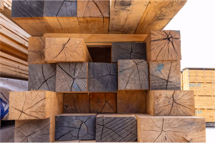 A stack of wooden beams showcasing different shades of timber, displayed in a lumber yard. The beams feature visible growth rings and grain patterns, with some showing signs of aging. The background includes additional stacks of lumber.