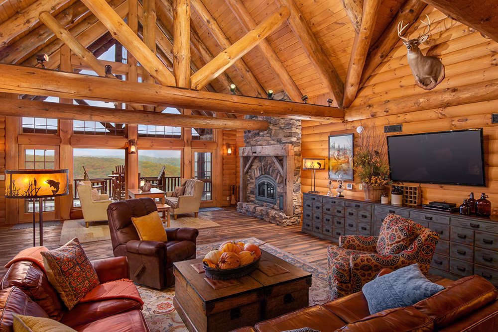 Log home great room featuring an exposed timber cathedral ceiling, stone wood-burning fireplace, large windows, and warm wood interiors.