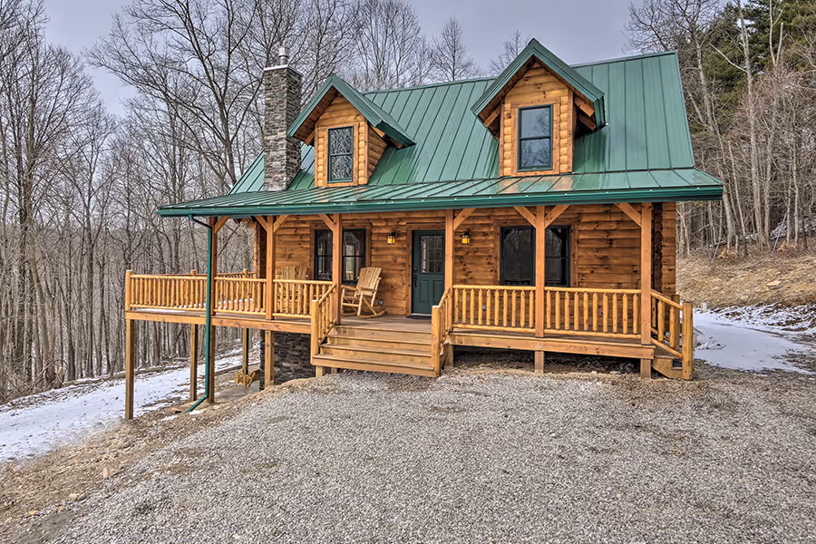A cozy wooden cabin with a green metal roof, featuring a porch with rocking chairs. It is surrounded by trees and a gravel driveway, with patches of snow on the ground, indicating a tranquil countryside setting.