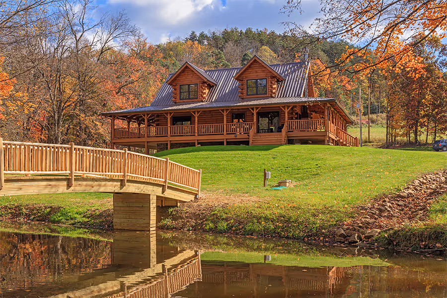 A spacious wooden lodge with a metal roof, situated by a tranquil pond. A wooden bridge leads to the house, surrounded by vibrant autumn foliage in various shades of orange and yellow.