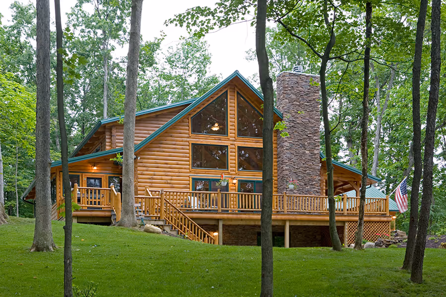 A large log cabin surrounded by lush green trees, featuring large windows and a stone chimney. A wooden deck wraps around the front of the cabin, with an American flag displayed. The landscaping includes well-maintained grass and natural elements.