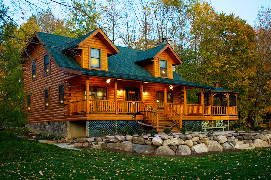 A charming log cabin nestled among colorful autumn trees, featuring a green roof and wooden porch. Stone pathways lead to the entrance, blending rustic elegance with natural surroundings.