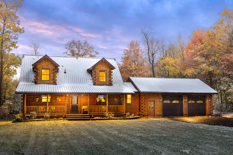 A charming log cabin with a metal roof surrounded by autumn trees, showcasing warm lights from the windows. The scene features a grassy area lightly dusted with snow, set against a colorful sunset sky.