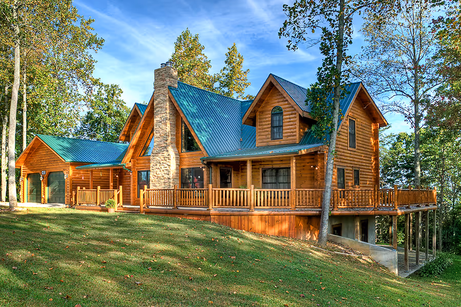 A large wooden lake house with a steep blue roof, surrounded by lush green trees. Features a spacious deck and stone chimney, set on a grassy slope. The warm wood tones and natural surroundings create a serene atmosphere.