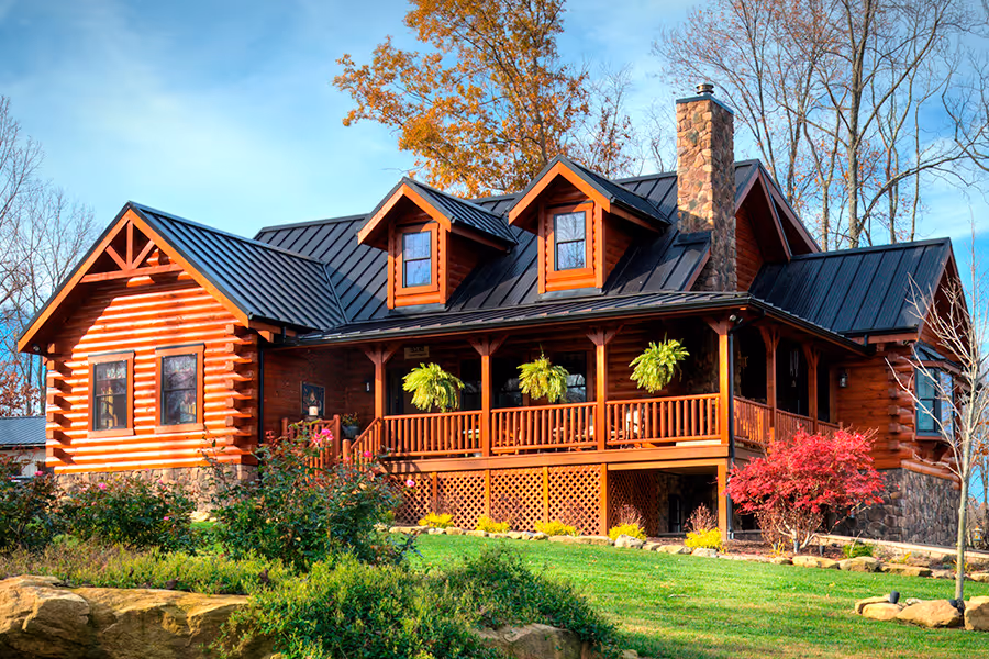 A charming log cabin with a dark metal roof, featuring a spacious front porch adorned with hanging ferns. The home is surrounded by lush green grass and vibrant autumn-colored trees, creating an inviting outdoor setting.