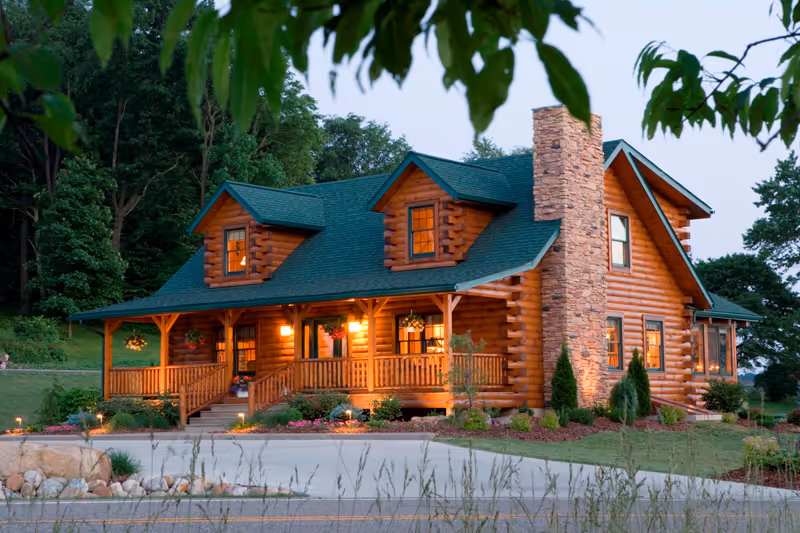 A charming log cabin with a green roof and stone chimney, surrounded by trees. The warm lights emanate from the porch and windows, enhancing the cozy ambiance of the home. A gravel driveway leads up to the house, with neatly landscaped gardens.