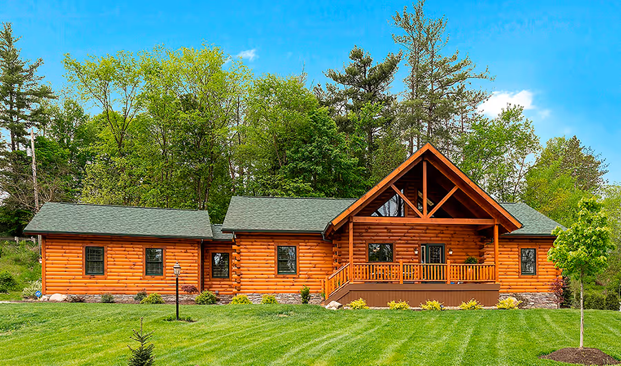 A charming log cabin with a large front porch, featuring a gabled roof and warm wooden exterior, surrounded by lush greenery and trees under a clear blue sky.