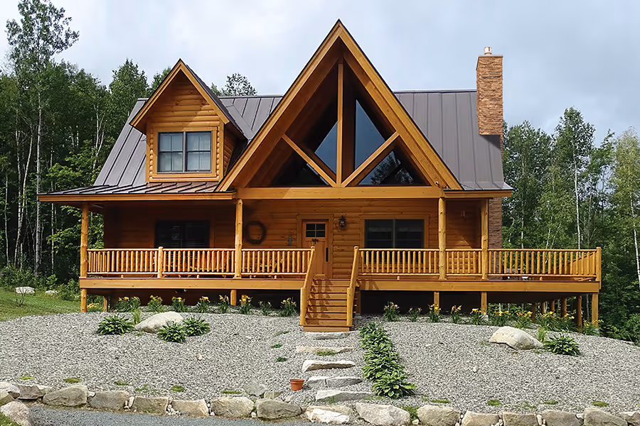 A rustic wooden cabin with a triangular roof, large front windows, and a spacious porch. The house is surrounded by greenery and rocky landscaping, set against a backdrop of trees under a cloudy sky.