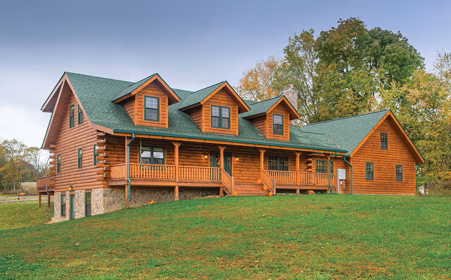 A large log cabin with a green roof, featuring multiple gabled sections and a spacious porch, stands on a grassy lawn surrounded by trees. The rustic design highlights wood textures and friendly architecture.