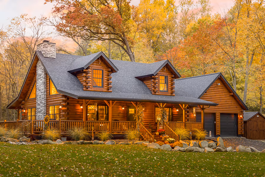 A charming log cabin surrounded by colorful autumn trees, featuring a welcoming porch, stone chimney, and warm glowing lights from the windows. A well-maintained lawn with a stone pathway leads to the entrance.
