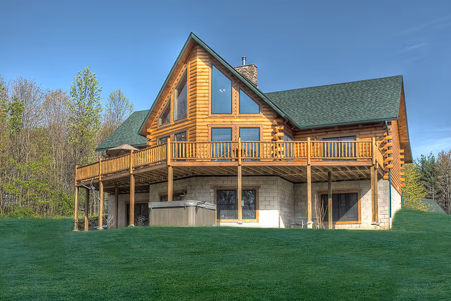 A spacious log cabin with a steeply sloped roof and large windows, surrounded by green trees. The cabin features a wide deck with wooden railings overlooking a well-manicured lawn, creating a welcoming outdoor space.