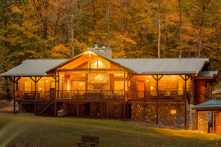 A cozy cabin nestled in a forest with autumn foliage, featuring a stone facade, large windows illuminating the interior, and a wraparound porch. Golden and orange leaves enhance the serene atmosphere of the wooded surroundings.