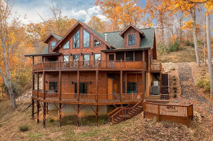 A large wooden cabin nestled in a forest, surrounded by vibrant autumn foliage. The house features multiple balconies and large windows, showcasing a blend of rustic charm and modern design. A winding path leads up to the structure from the lower level.
