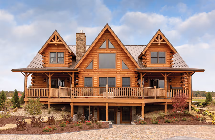 A large log cabin with a wooden deck, featuring multiple gabled roofs and large windows. The cabin is surrounded by landscaped gardens and trees, offering a rustic yet modern appearance against a cloudy sky.