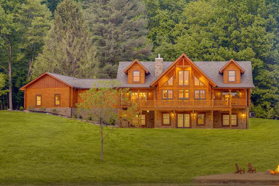A large wooden cabin-style house with multiple gables and expansive balconies, surrounded by lush green grass and trees. The house has warm lights glowing from its windows, creating a welcoming ambiance in the evening light.