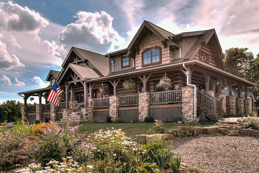 A large, rustic-style house with stone and wood elements, featuring a deck with wooden railings, surrounded by vibrant flowers. An American flag waves in the front yard under a blue sky filled with fluffy clouds.