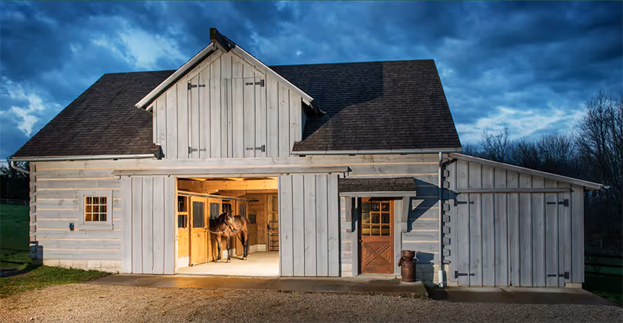 A rustic wooden barn with an open front, illuminated warmly from inside. Two horses stand near the entrance, framed by a cloudy twilight sky. The barn features a peaked roof and a weathered appearance, set in a grassy landscape.