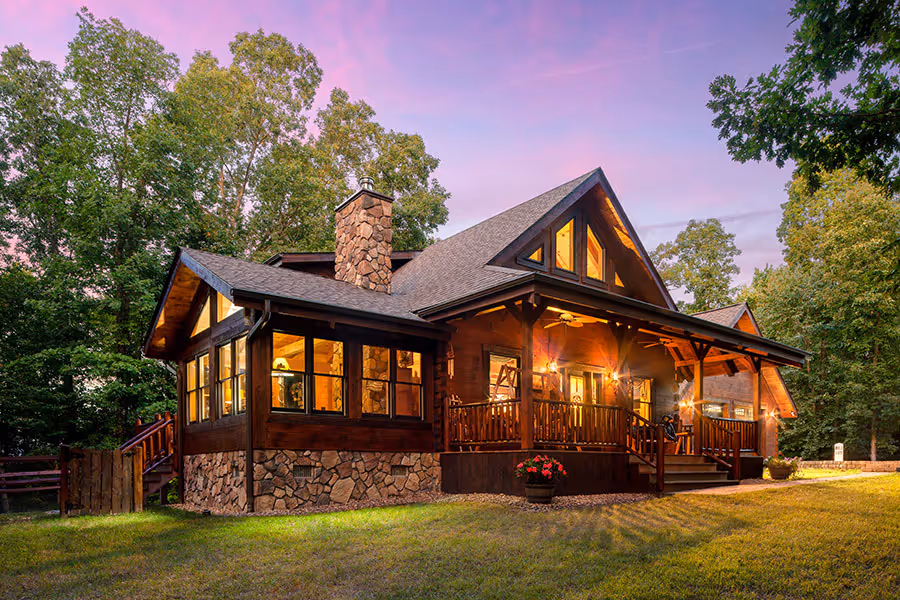 A charming wooden cottage with a stone facade and large windows, surrounded by tall trees and a grassy area, illuminated by a warm evening glow under a colorful sky.