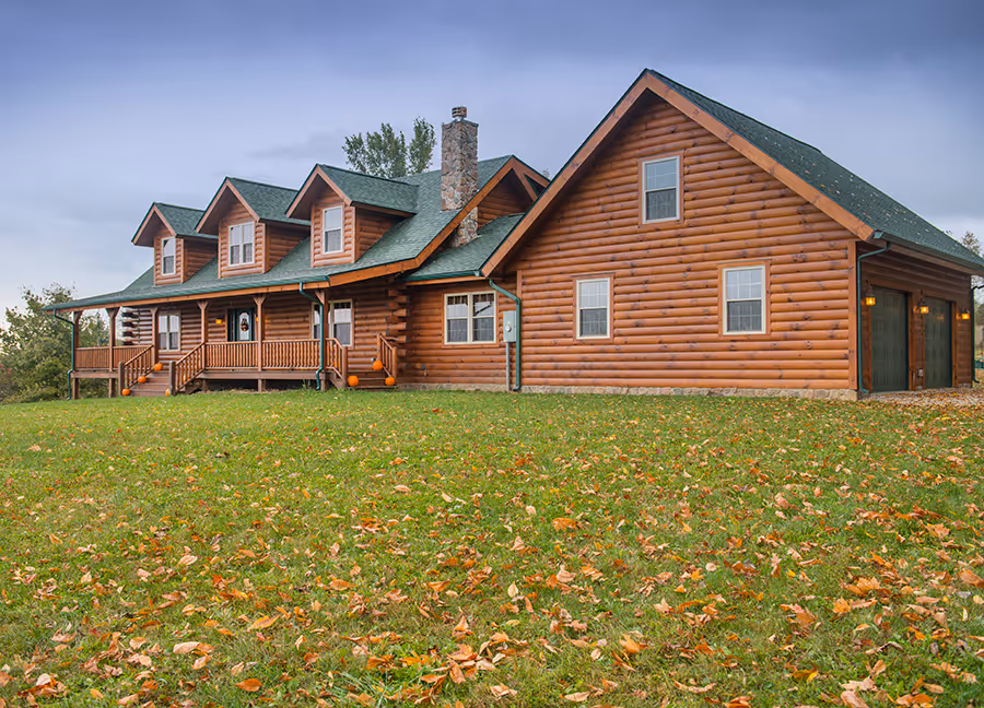 A spacious log cabin surrounded by autumn foliage, featuring a green roof and a large porch with multiple entrances. The cabin has windows and wooden siding, set against a cloudy sky.
