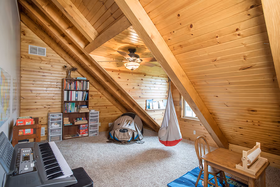 A cozy attic room with wooden sloped ceilings, featuring a small desk and chair, a keyboard, and a colorful rug. A tent for children is set up in the corner beside a bookshelf filled with books. Natural light streams in from a window.