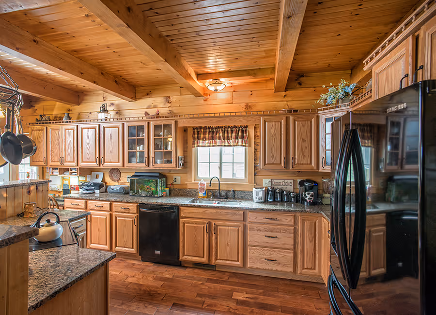 A rustic kitchen featuring wooden cabinets and exposed beams, with a granite countertop. The kitchen includes a black refrigerator, a stainless steel sink, and a small fish tank on the counter, complemented by warm wooden tones.