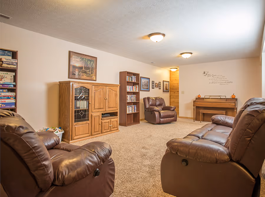 A cozy living room featuring two reclining chairs and a wooden entertainment center. A bookshelf filled with DVDs is against the wall, with a piano and decorative accents in the space. Warm lighting creates a welcoming atmosphere.