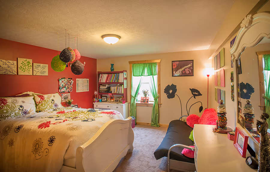 A cozy bedroom featuring a white bed with colorful floral bedding. The walls are painted in deep red and light beige, adorned with framed artworks. A green curtain hangs by the window, and colorful hanging decorations add vibrancy.