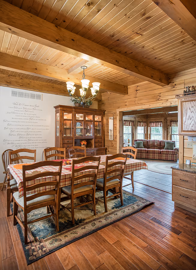Rustic dining room with wooden beams and a chandelier above a wooden table with eight chairs. A china cabinet displays dishes. A cozy seating area is visible through an open doorway. Earthy tones and a welcoming atmosphere enhance the space.