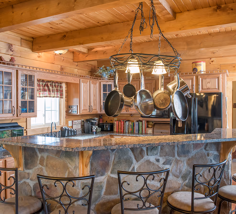 A rustic kitchen featuring wooden beams and cabinets, with a central island that has a stone base. Hanging from the ceiling are several pots and pans. The background displays shelves filled with cookbooks.