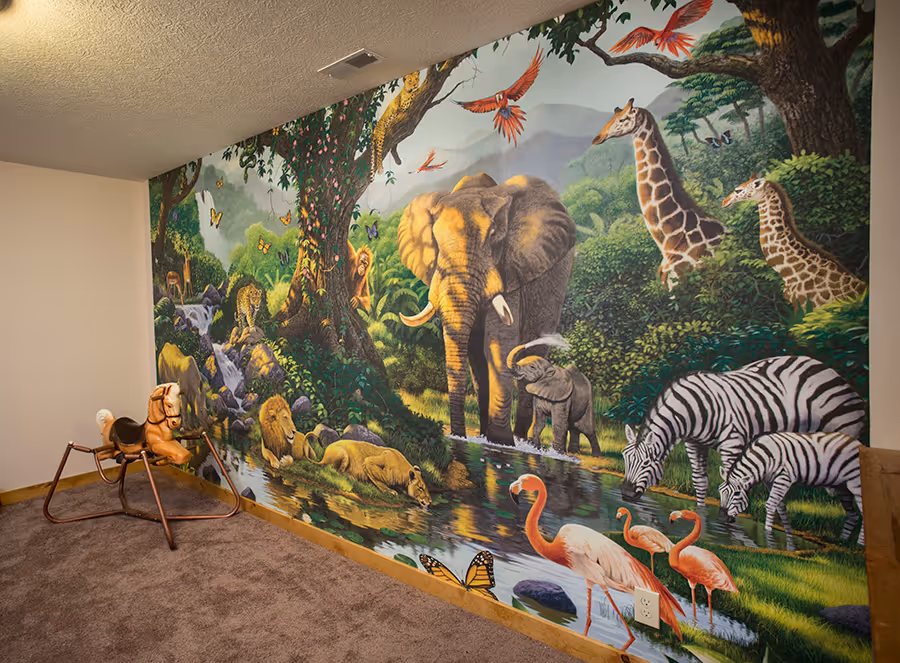 A vibrant mural depicting a jungle scene featuring various animals including elephants, zebras, giraffes, and flamingos, with lush greenery and a serene river. A wooden rocking horse is positioned in the foreground on a carpeted floor.