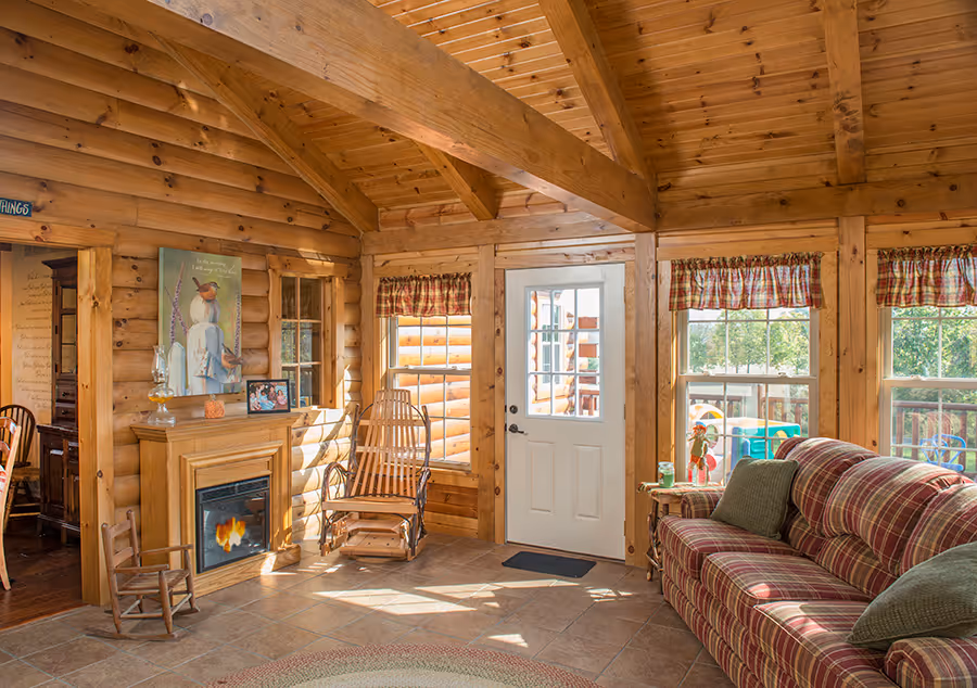 A cozy living room with wooden walls and a high ceiling, featuring a striped sofa, a small fireplace, and a door leading to an outdoor area. Natural light fills the room through large windows adorned with curtains.
